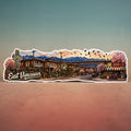 A die-cut sticker featuring a colorful illustrated scene of East Vancouver, with buildings, a train, mountains, cherry blossoms, and a sunset sky, shown standing upright against a gradient background.